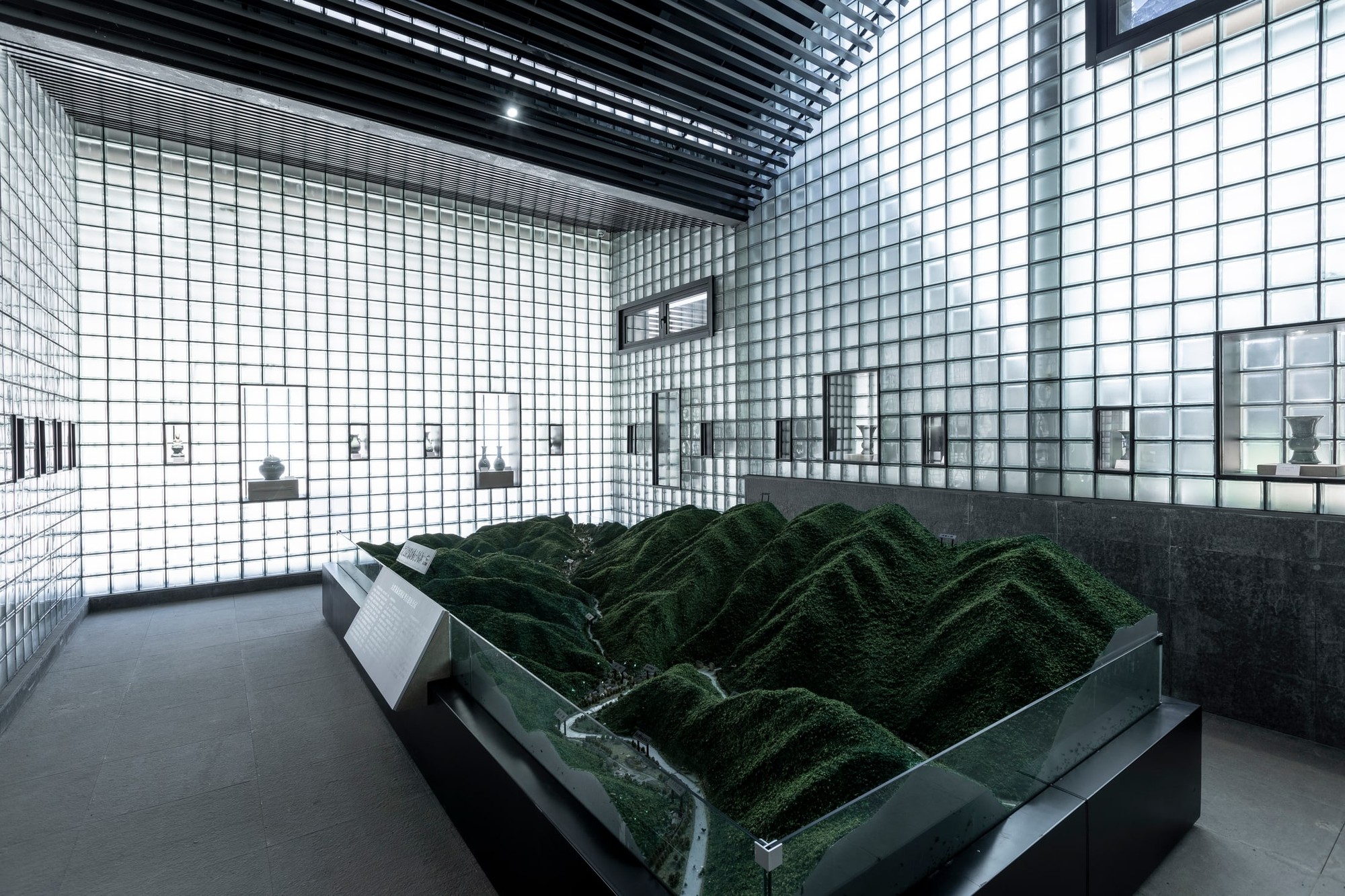 A physical landscape model forms a central exhibit within the glass-block enclosure.